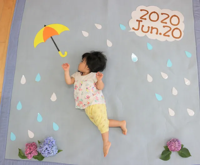 寝相アートで傘と雨つぶの絵の上に寝ころぶ赤ちゃん